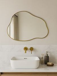 Organic-shaped gold-framed mirror hung above a white vessel sink in a neutral bathroom, reflecting curtains and natural light