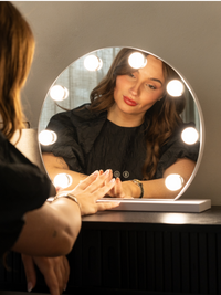 Woman using white round Hollywood vanity mirror with 9 lit LED bulbs, touching the touch controls on the base, reflection…