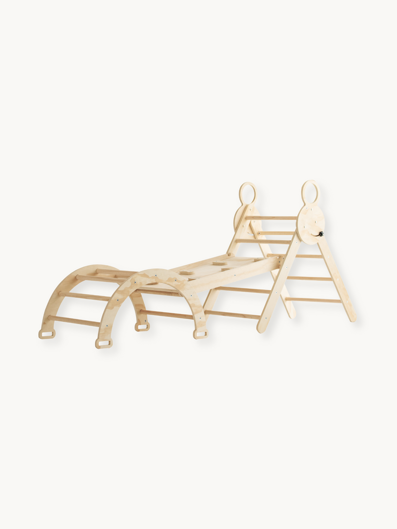 Natural wood Pikler-style climbing set with arch rocker, triangle ladder, and ramp slide connected, angled view on white b…
