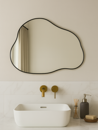Organic-shaped Matteo mirror with black PVC frame hung above a white vessel sink and brass wall tap in a minimal bathroom…