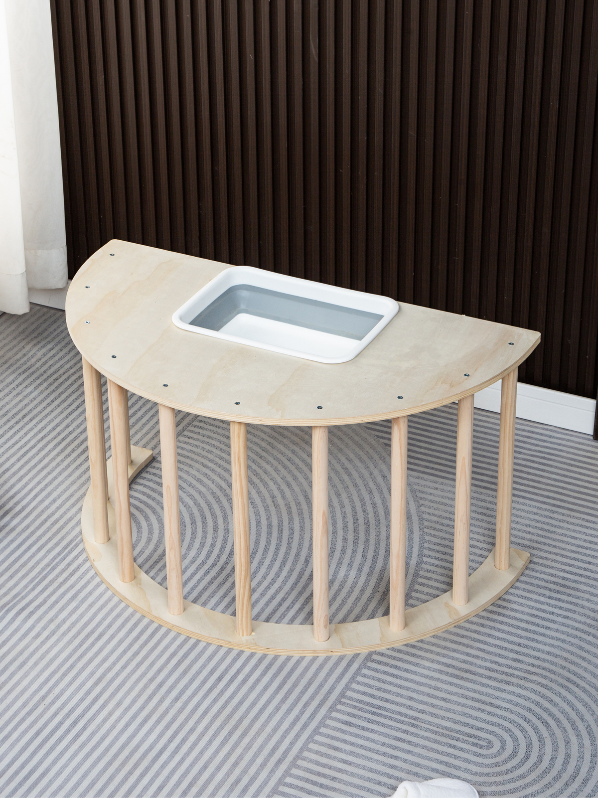 Natural wood semicircular play table with spindle legs, viewed from above at an angle, grey plastic storage bin recessed i…
