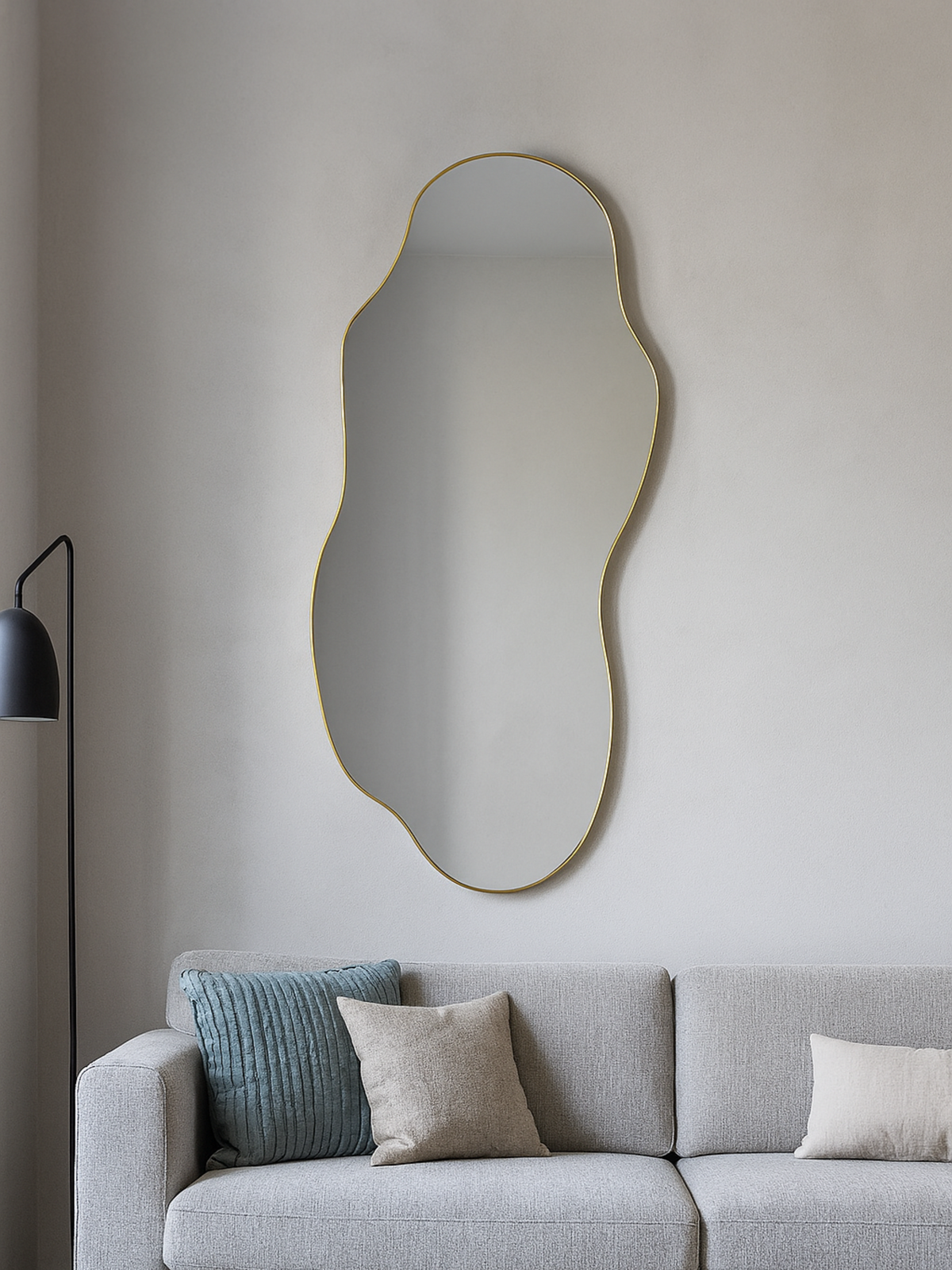 Luciano organic mirror with thin gold PVC frame hung above a grey sofa in a living room, reflecting soft ambient light