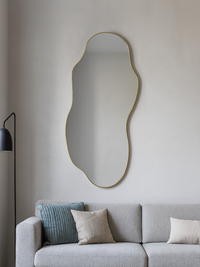 Luciano organic mirror with thin gold PVC frame hung above a grey sofa in a living room, reflecting soft ambient light