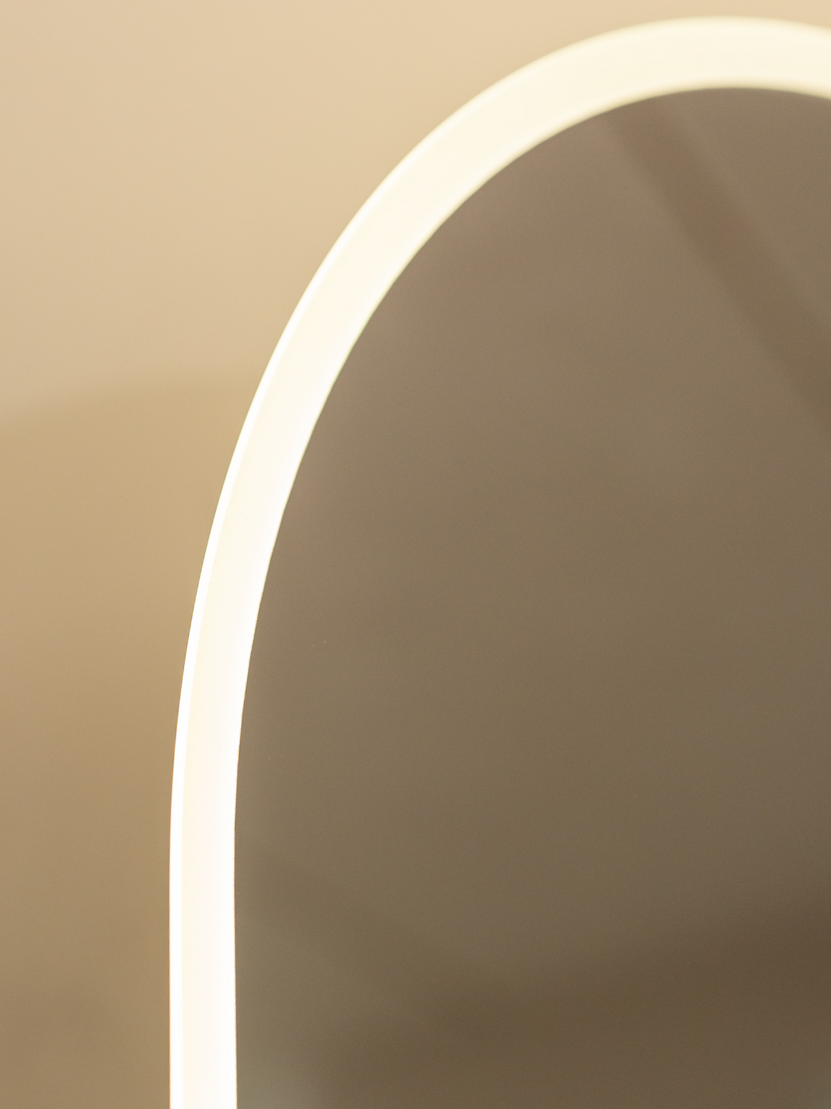 Close-up of glowing warm white LED perimeter strip along left curve of oval frameless standing mirror against beige wall