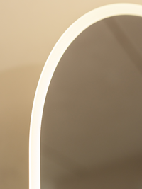 Close-up of glowing warm white LED perimeter strip along left curve of oval frameless standing mirror against beige wall