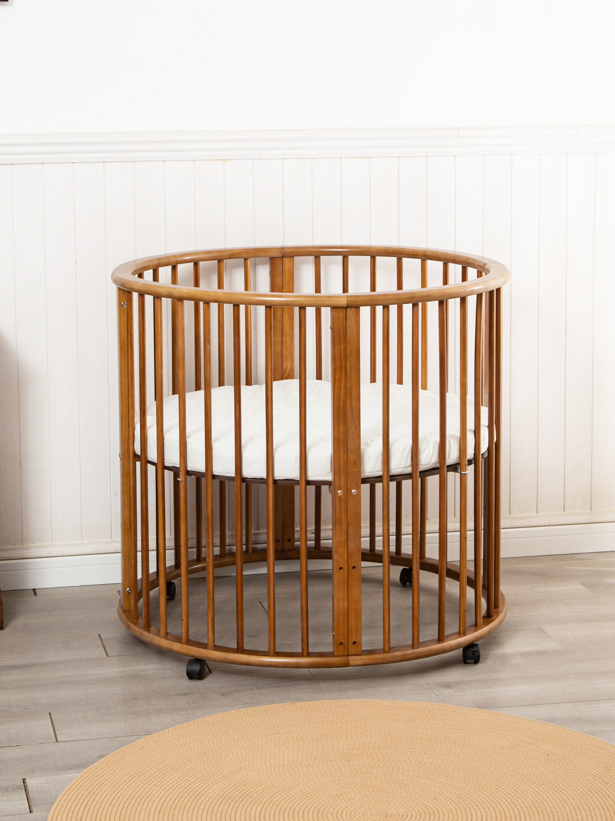Brown round wooden baby playpen with slatted sides and white mattress, on castors, in a nursery with light wood floor and…