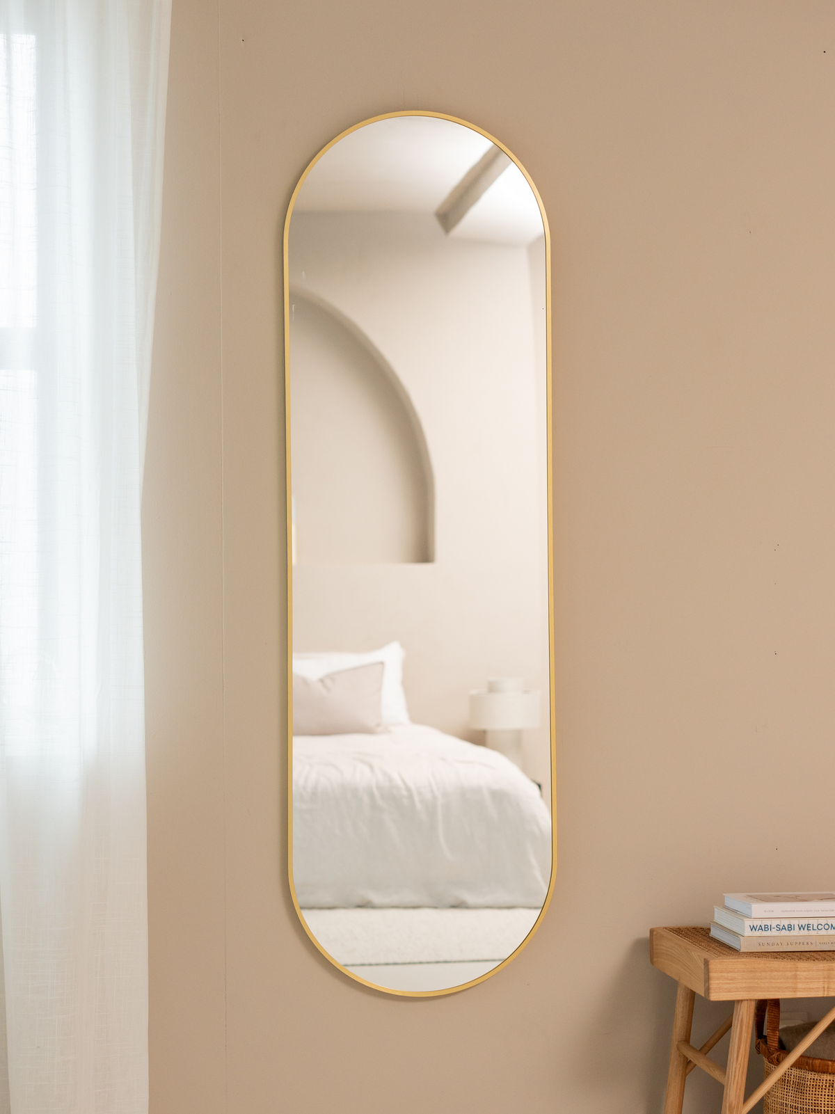 Tall gold-framed capsule mirror hung on a warm beige bedroom wall, reflecting a white bed and arched headboard, rattan sto…