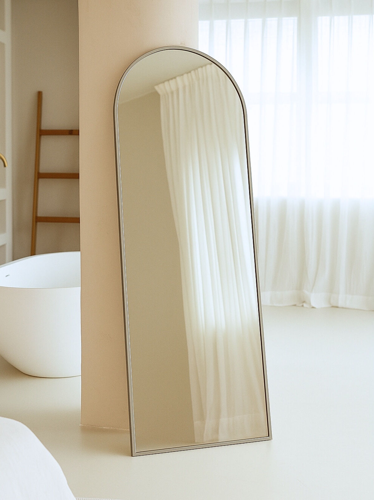 Taupe arched full-length mirror leaning against a beige wall in a bright bathroom, freestanding angle, white curtains refl…