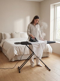 Woman ironing a white shirt with a steam iron on a black ironing board in a sunlit neutral bedroom, steam visibly rising