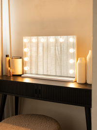 White Hollywood makeup mirror with 12 glowing LED bulbs on a dark dressing table, styled with white vases and beauty produ…