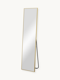 Full-length Angelo mirror with slim gold frame leaning at an angle, black metal floor stand visible from the side, white b…
