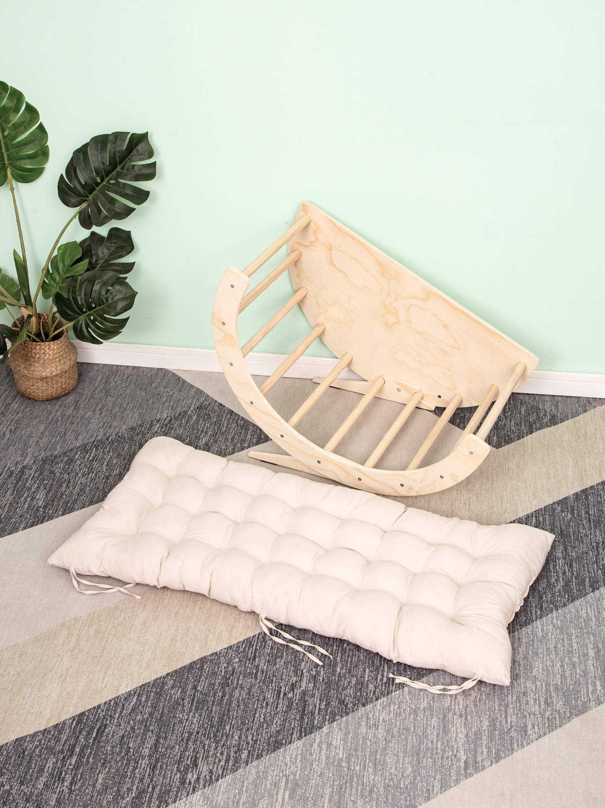 Natural birch climbing arch tilted on its side beside a cream tufted cushion with tie strings, on grey striped carpet