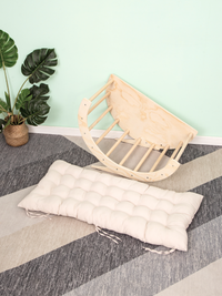 Natural birch climbing arch tilted on its side beside a cream tufted cushion with tie strings, on grey striped carpet