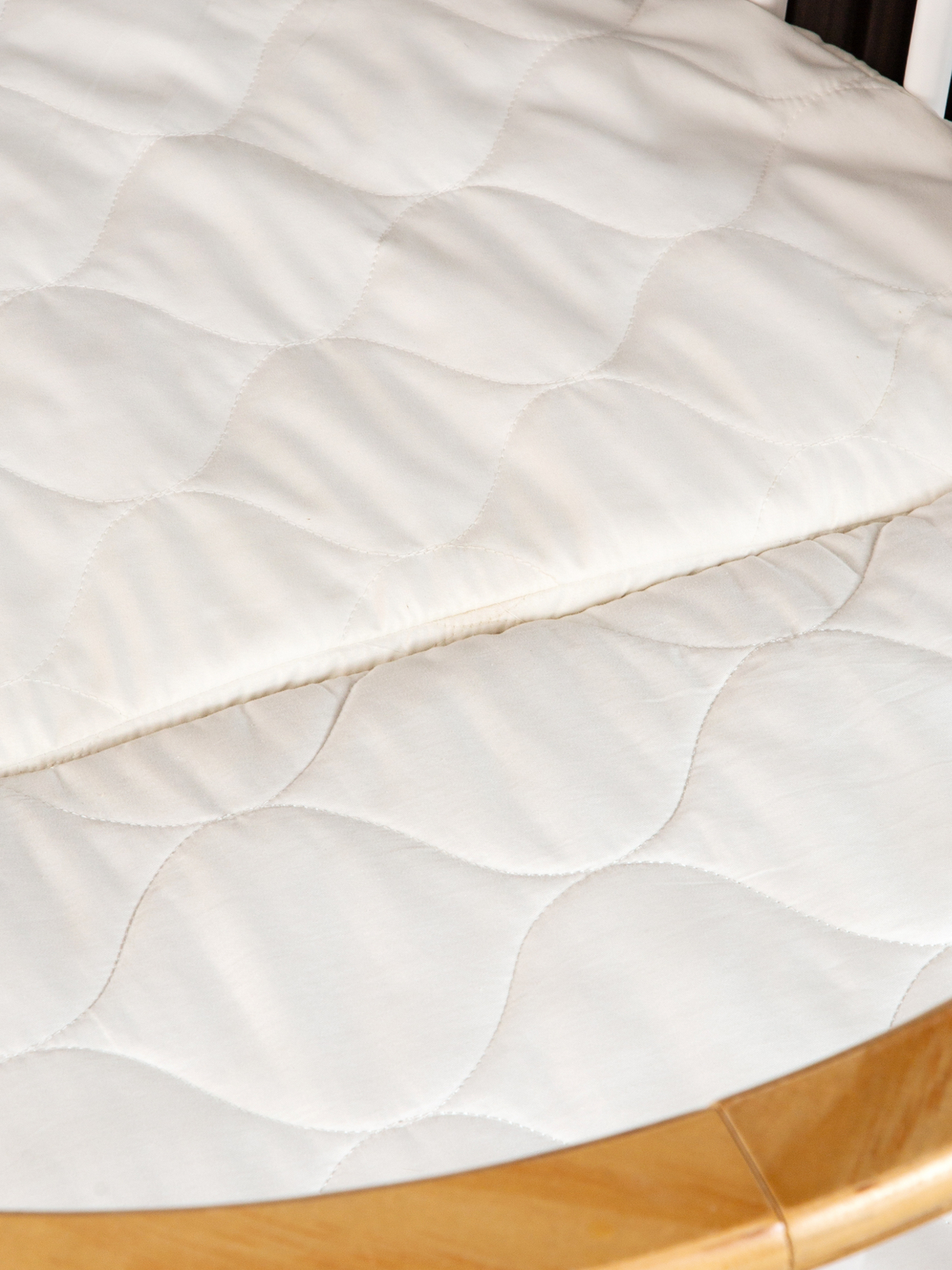 Close-up of white quilted mattress with scallop stitch pattern fitted inside round wooden playpen, natural wood rim visibl…