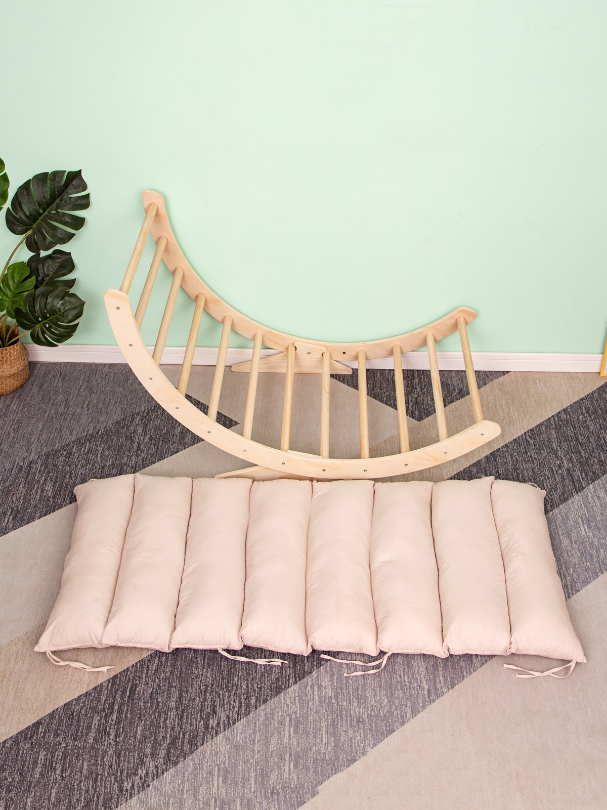Natural wood climbing arch rocker leaning against mint wall, blush pink segmented cushion laid flat on patterned floor, mo…