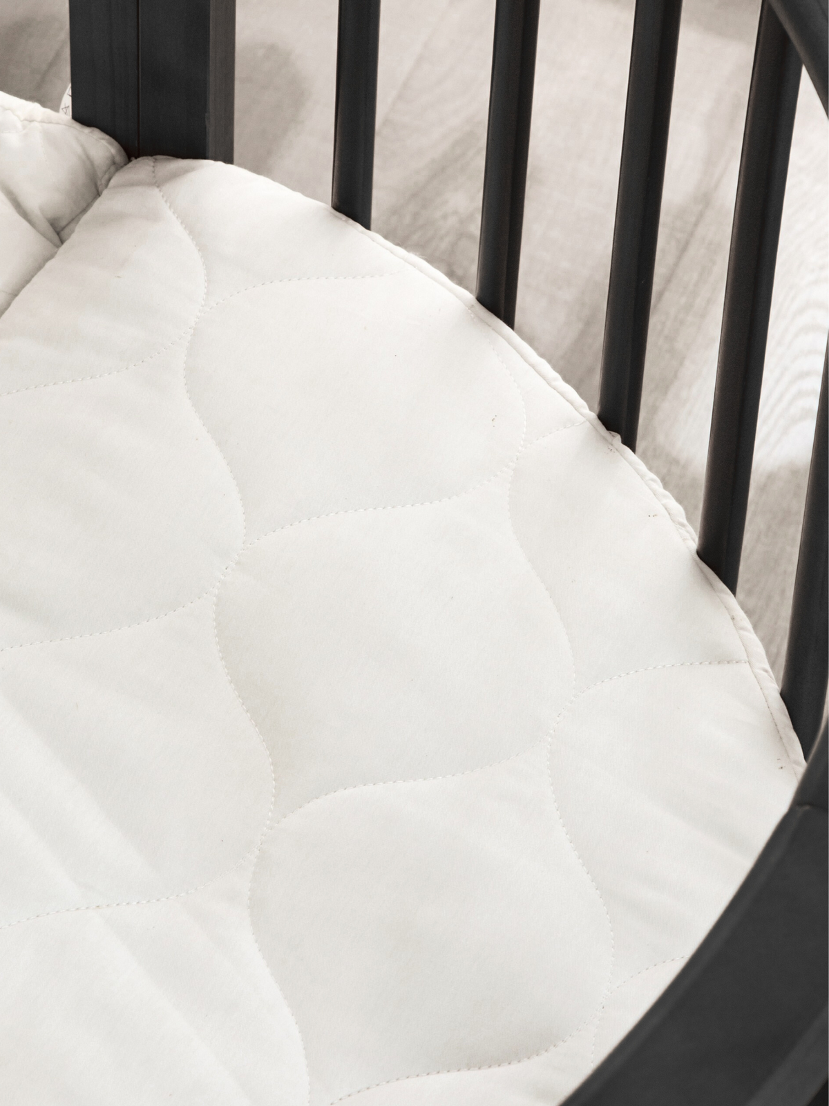 Close-up of white quilted oval mattress inside black round wooden playpen, showing curved stitching pattern and piped edge