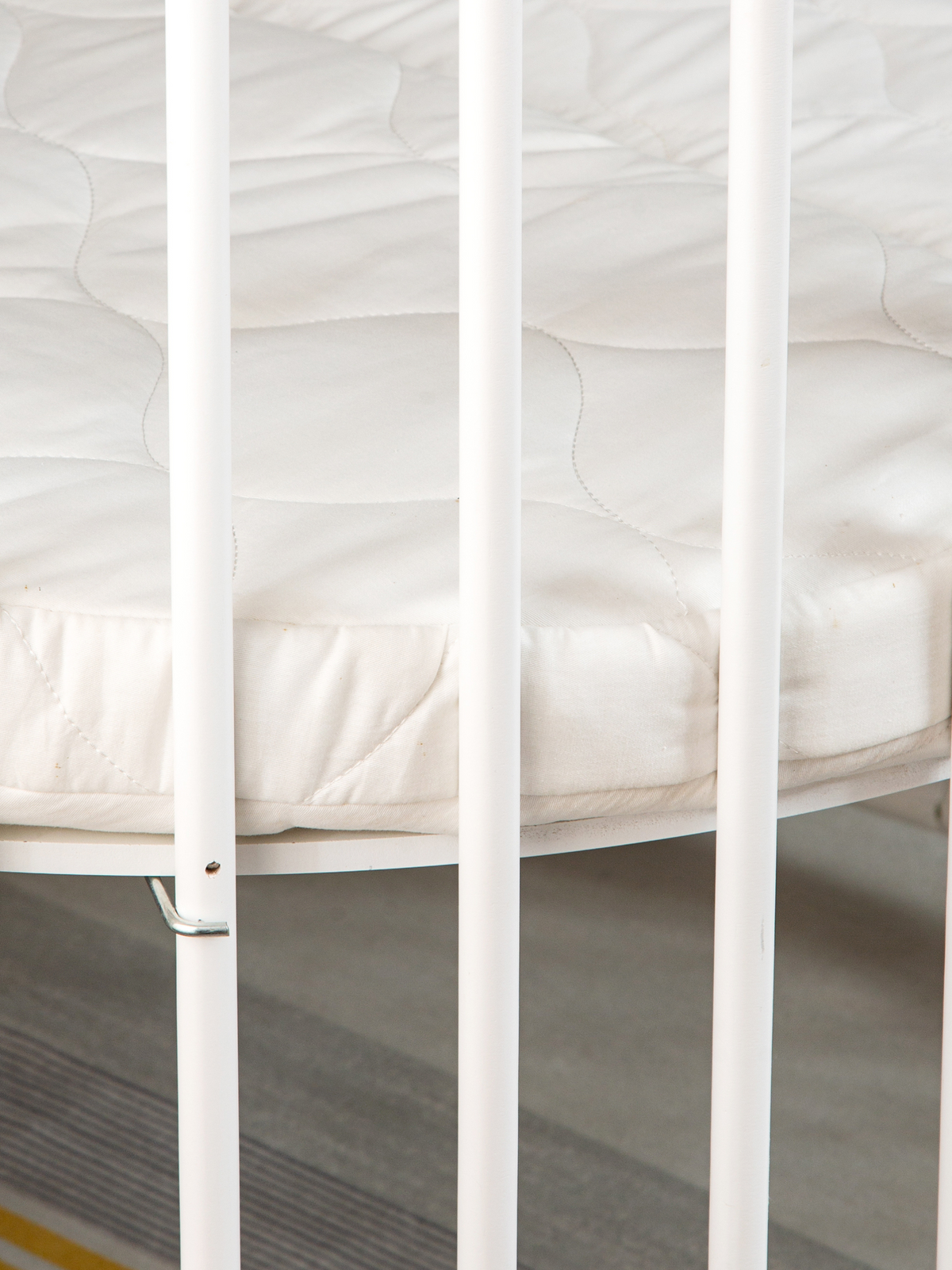 Close-up of white vertical slats on round playpen with quilted circular mattress visible, shot from ground-level angle