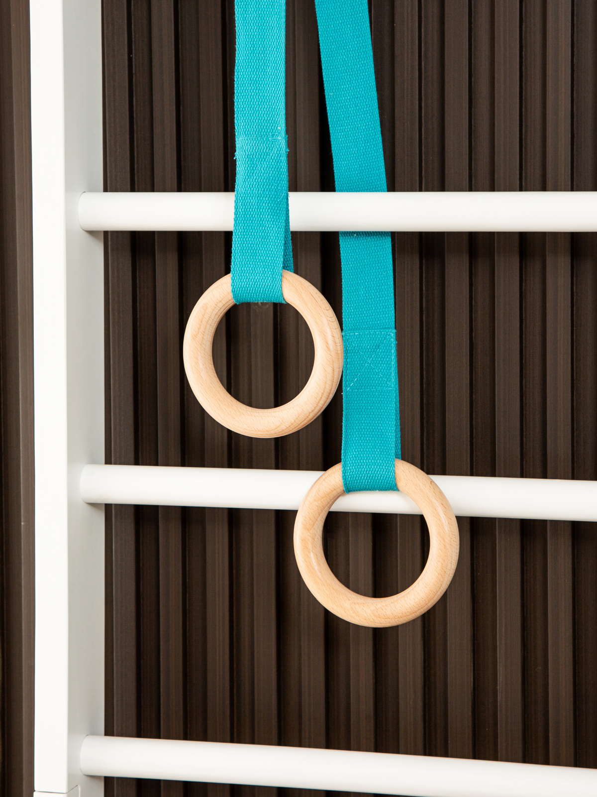 Two natural wood gymnastic rings on teal fabric straps, close-up detail against a dark slatted wall panel and white frame…