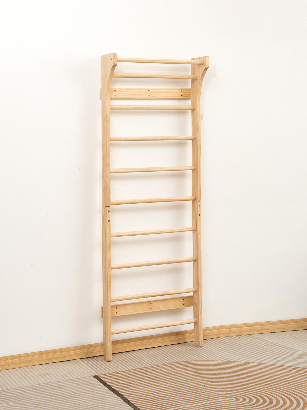 Natural wood Swedish wall bar leaning against white wall, full-length angled view on striped rug, showing two-section fram…