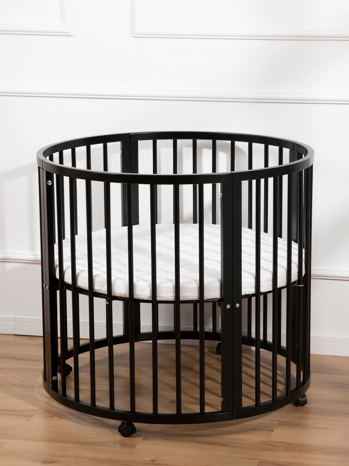Black round wooden playpen with vertical slats and white quilted oval mattress, shown on light wood floor against white pa…