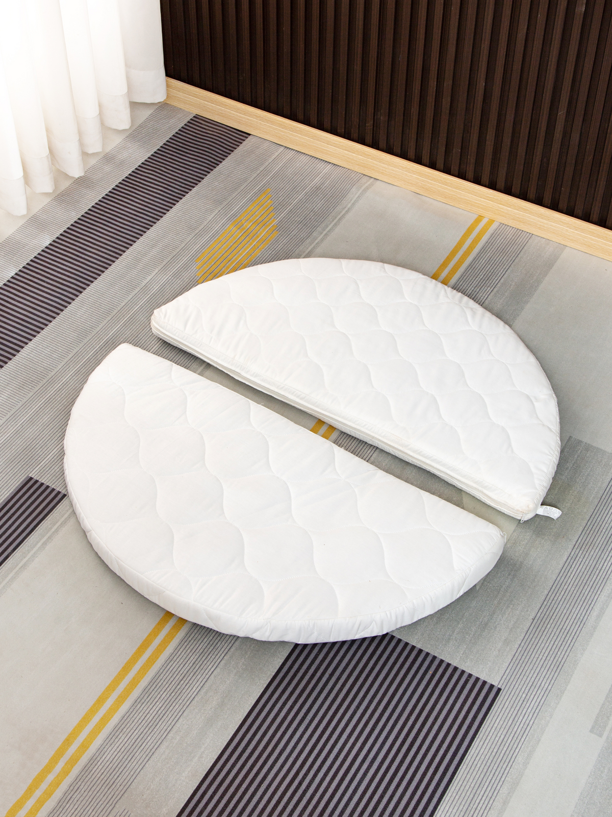 White quilted round mattress split into two half-circle sections, laid flat on a striped grey and yellow rug, overhead view.