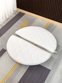 White quilted round mattress split into two half-circle sections, laid flat on a striped grey and yellow rug, overhead view.