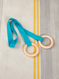 Two natural wood gymnastic rings on teal fabric straps laid flat on a grey and yellow striped textile surface