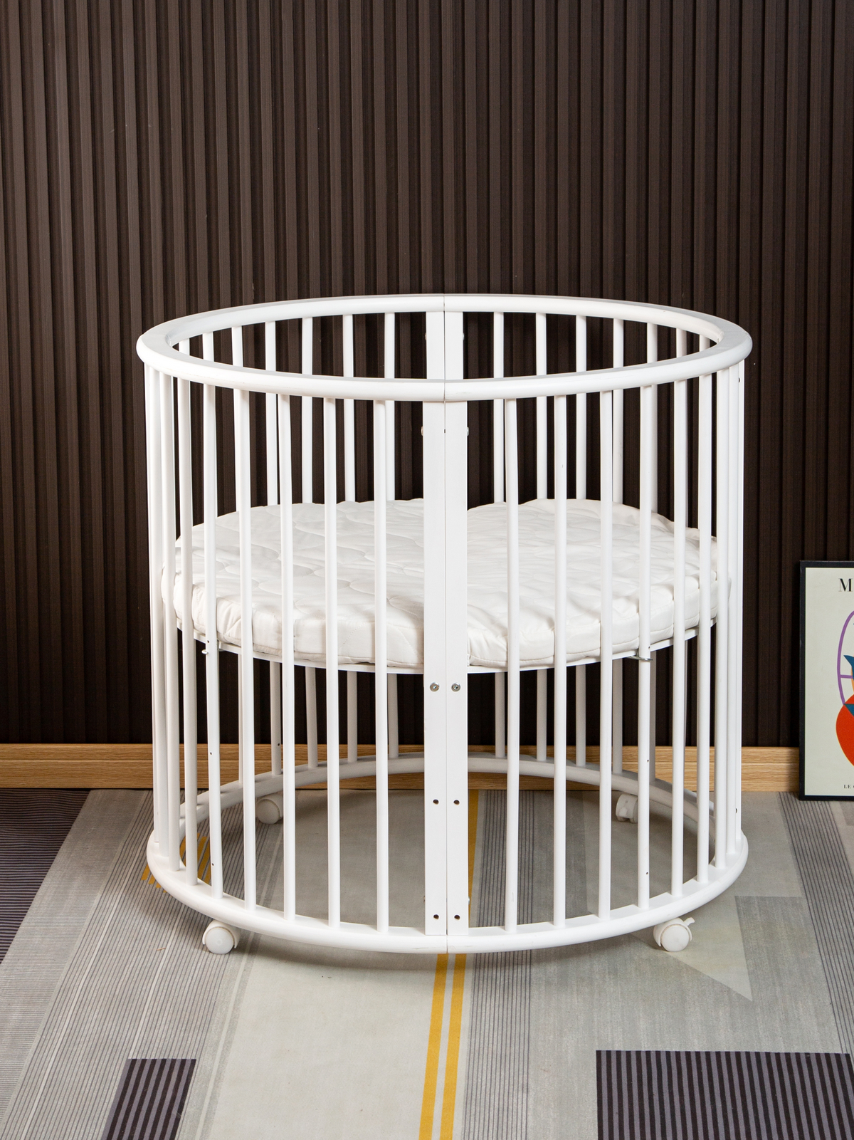White round wooden playpen with vertical slats and quilted circular mattress on casters, front angle, styled in modern roo…