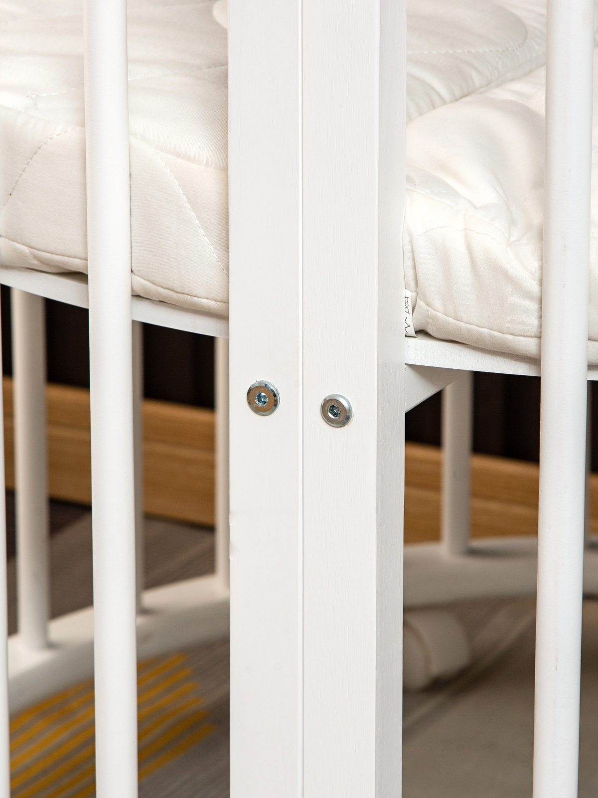 Close-up of two silver bolt fixings on white wooden playpen slat, with quilted cream mattress visible behind bars