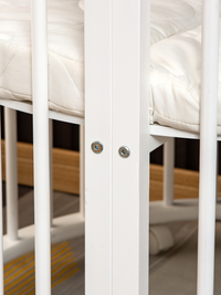 Close-up of two silver bolt fixings on white wooden playpen slat, with quilted cream mattress visible behind bars