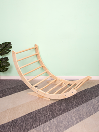 Natural wood climbing arch rocker leaning against mint wall without cushion, angled three-quarter view on striped carpet,…