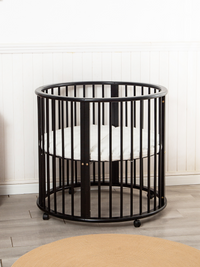 Black round wooden playpen on casters with white mattress inside, photographed front-on against white panelled wall with t…