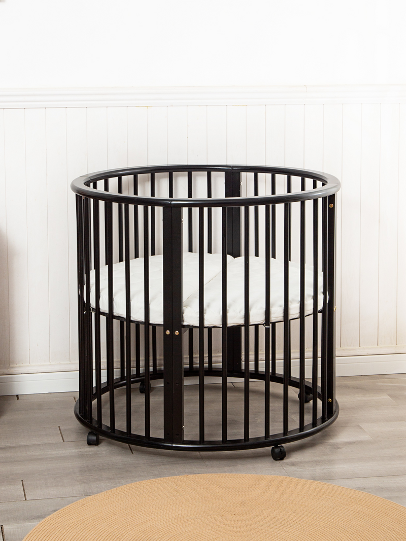 Black round wooden playpen on casters with white mattress inside, photographed front-on against white panelled wall with t…