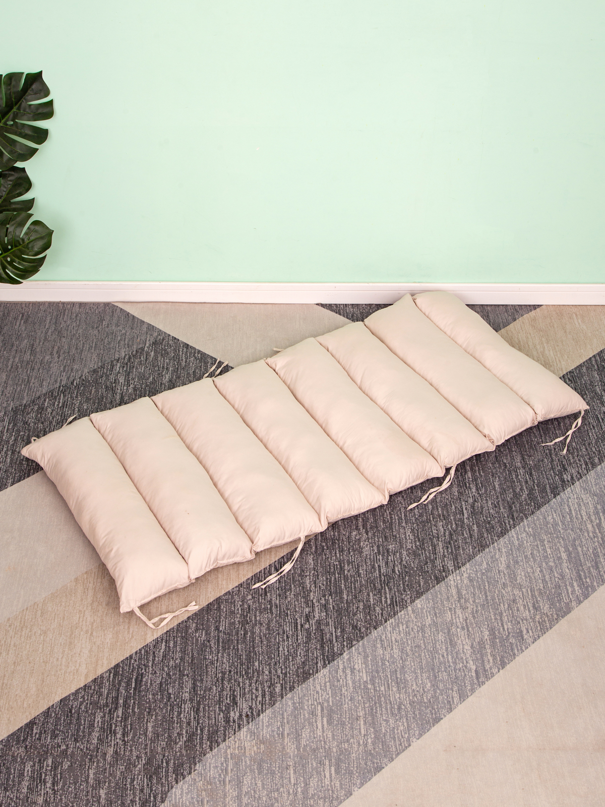 Cream quilted cushion laid flat on striped carpet, showing channel stitching and fabric ties along the sides, mint wall be…