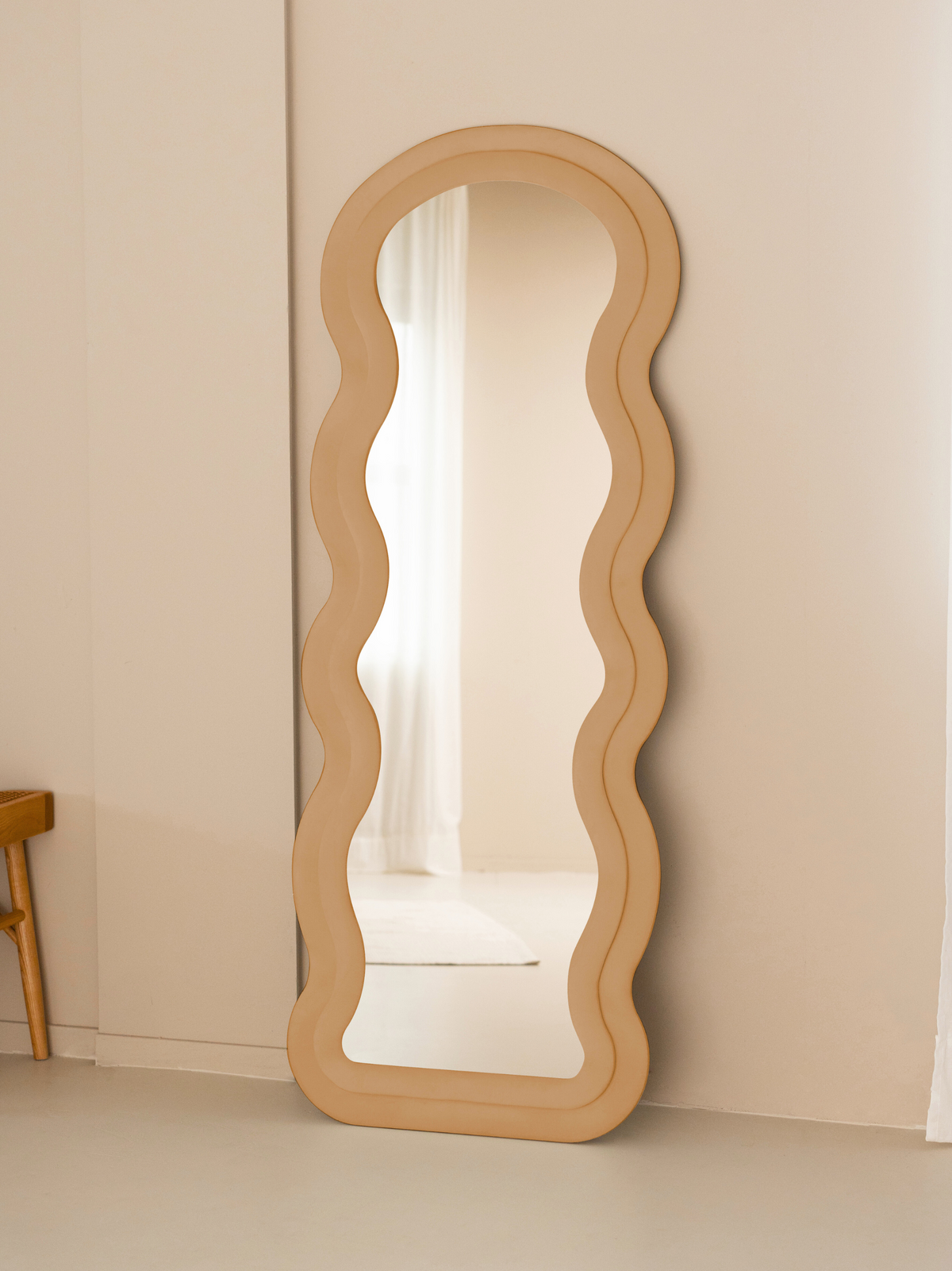 Taupe beige suede velvet wavy floor mirror leaning against cream wall beside a wooden chair, lifestyle setting