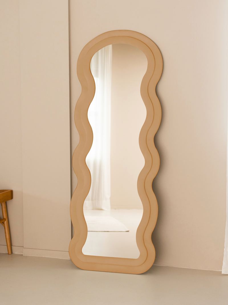 Taupe beige suede velvet wavy floor mirror leaning against cream wall beside a wooden chair, lifestyle setting