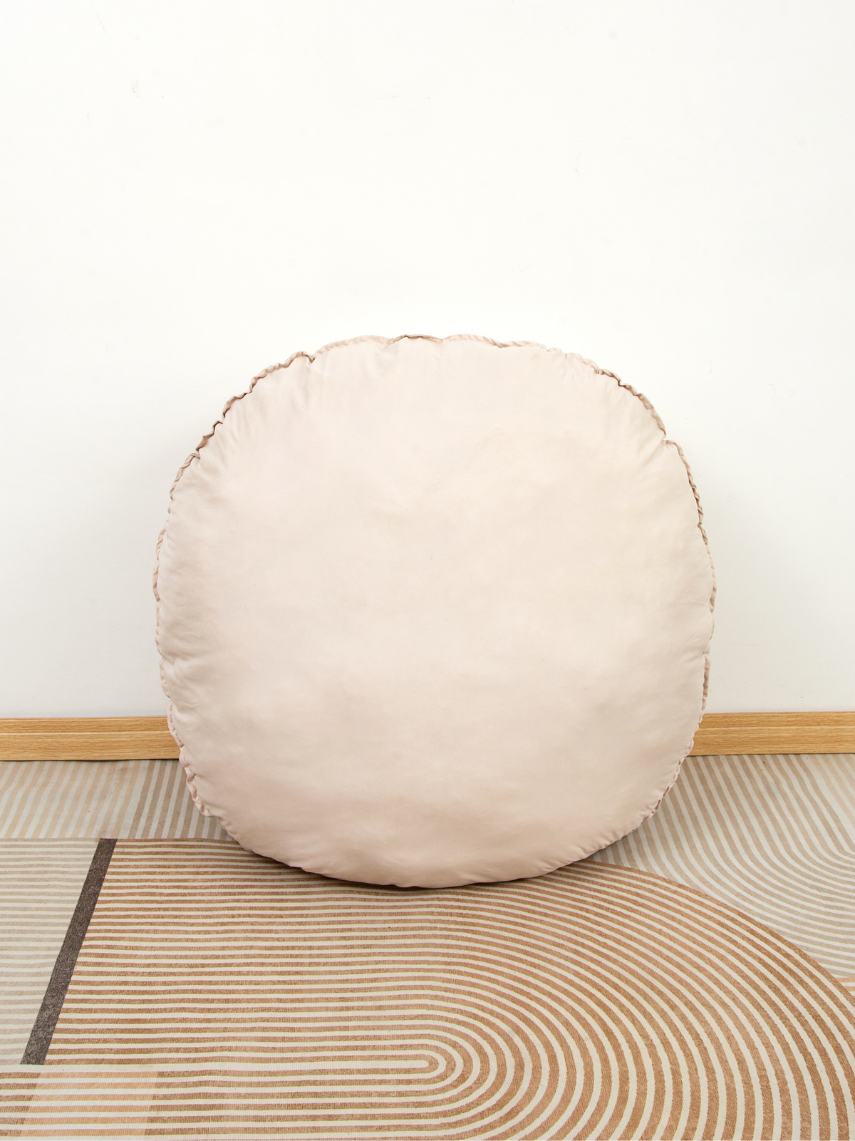 Blush-toned round floor cushion leaning against white wall on geometric striped rug, showing plain back panel and scallope…