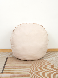 Blush-toned round floor cushion leaning against white wall on geometric striped rug, showing plain back panel and scallope…