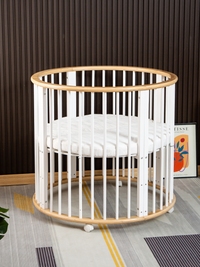 Round wooden playpen Sami with white slats and natural wood rim, white fitted mattress inside, on castors, styled in nurse…