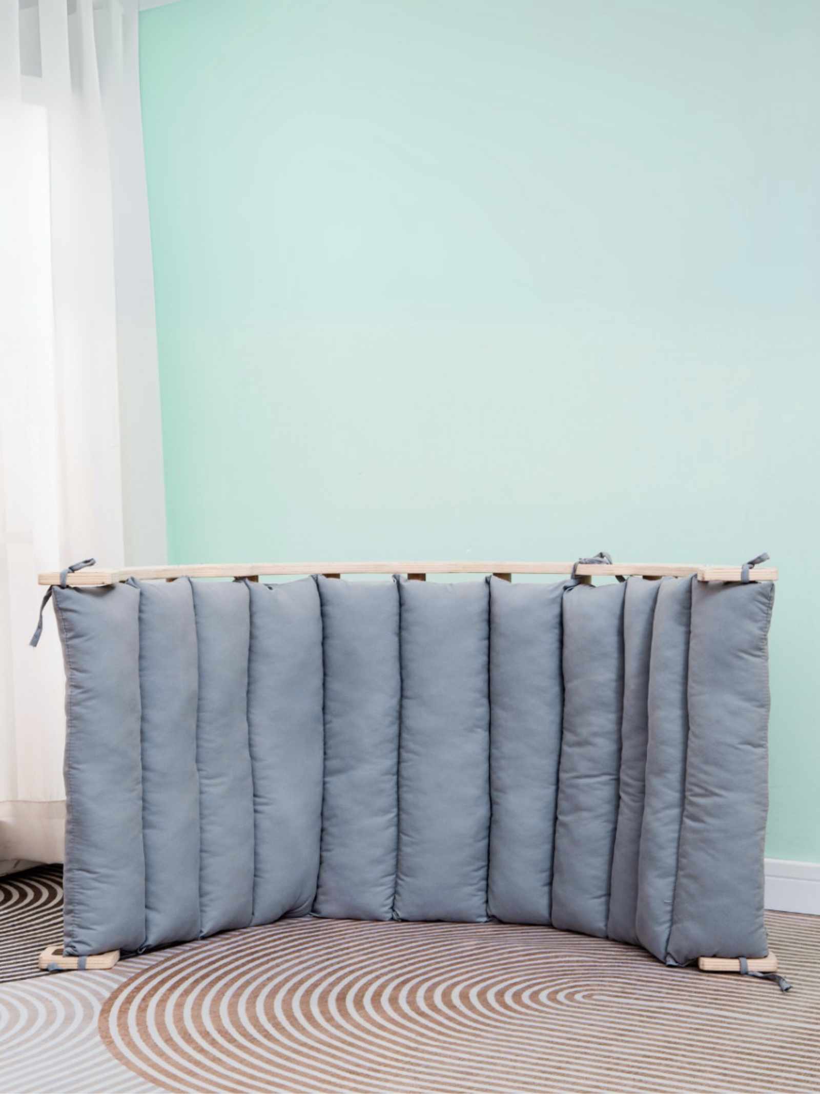 Grey quilted cushion with tie fastenings draped over a natural wood climbing arch rocker, lifestyle shot against mint wall
