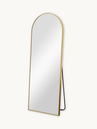 Angled three-quarter view of full-length arched mirror with slim gold aluminium frame and black metal easel stand, white b…