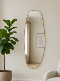 Tall asymmetric oval gold-framed mirror hung on cream wall beside fiddle-leaf fig plant and white armchair, lifestyle setting