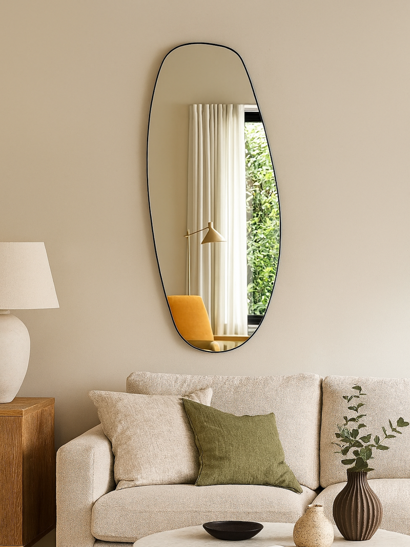 Leandro organic mirror with slim black frame hung above a cream sofa in a living room, reflecting curtains and greenery