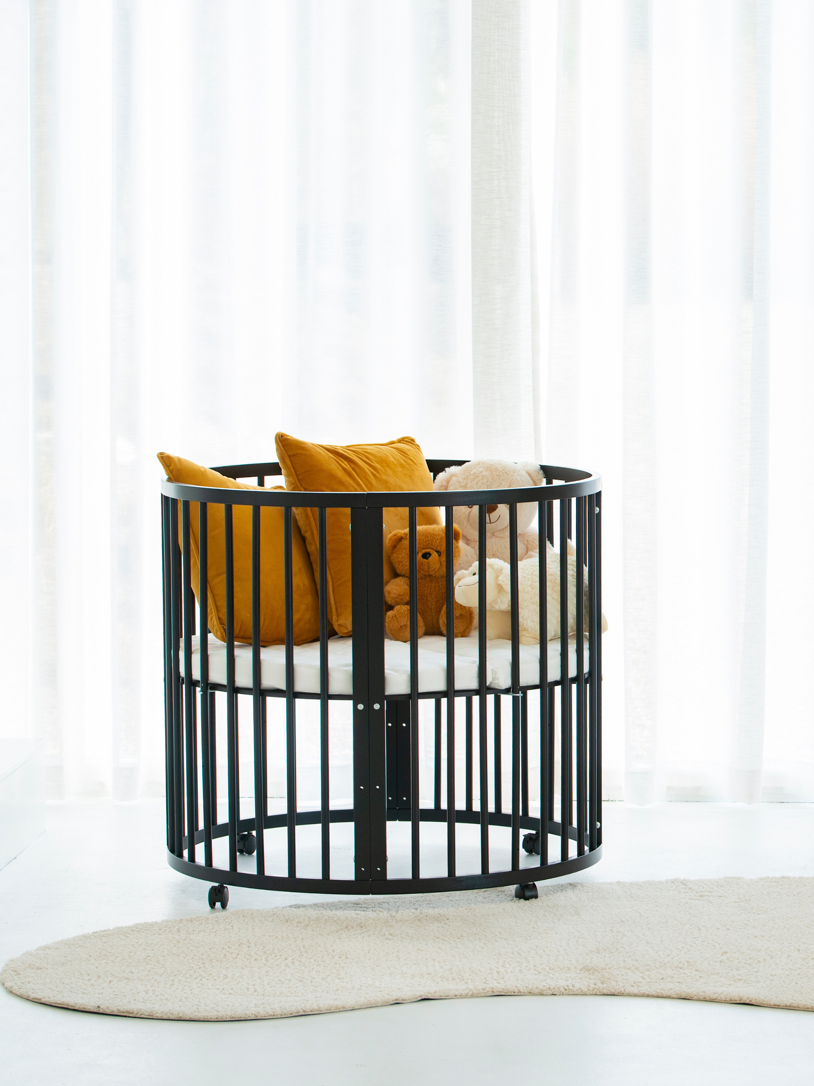 Black round wooden crib on castors styled with mustard cushions and plush teddy bears, bright nursery setting, side angle