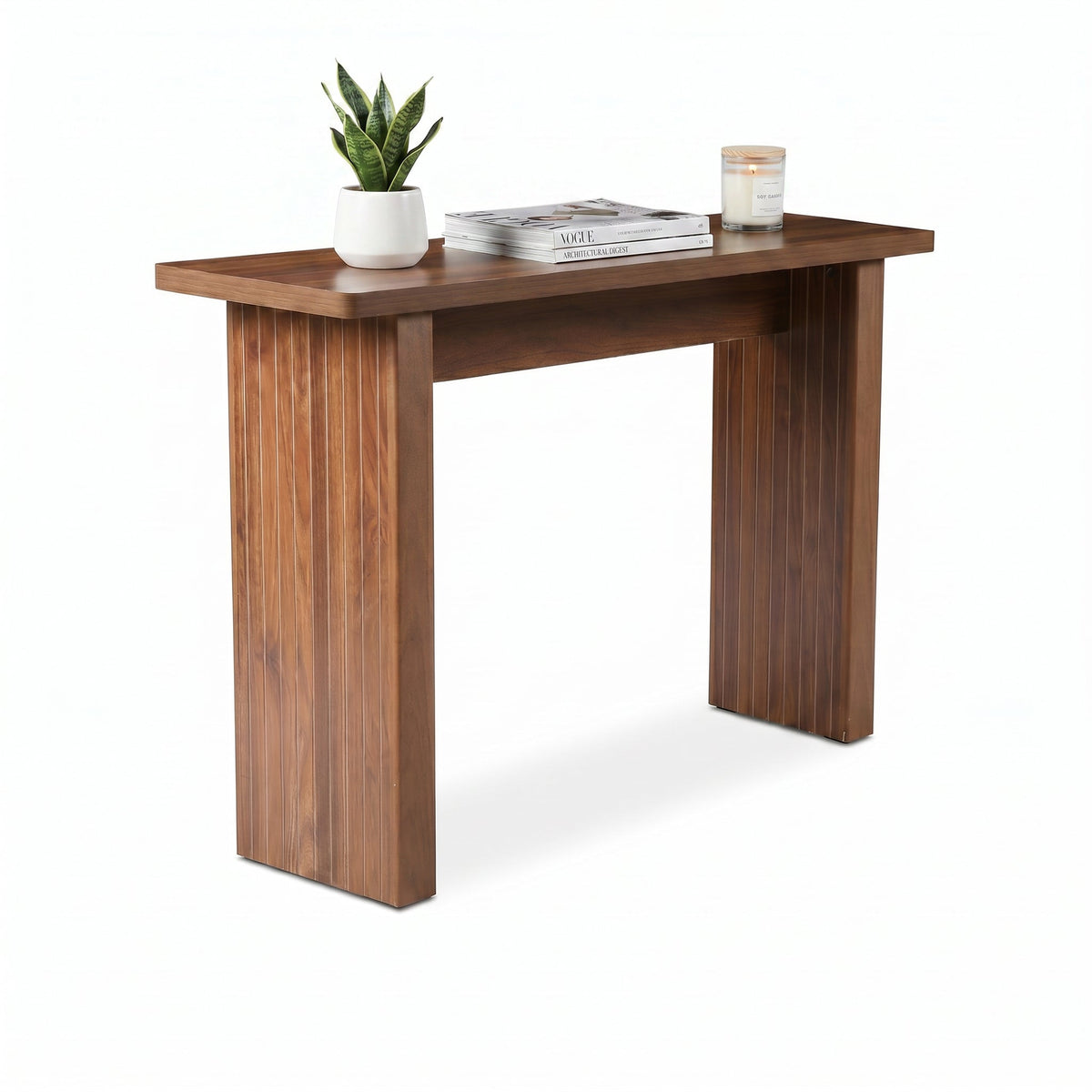 Walnut console table angled view styled with snake plant, stacked magazines, and lit candle on top, white background