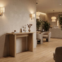 Narrow light oak console table styled in a warm spa-like interior, front-angle view, with framed art and dried floral vase…