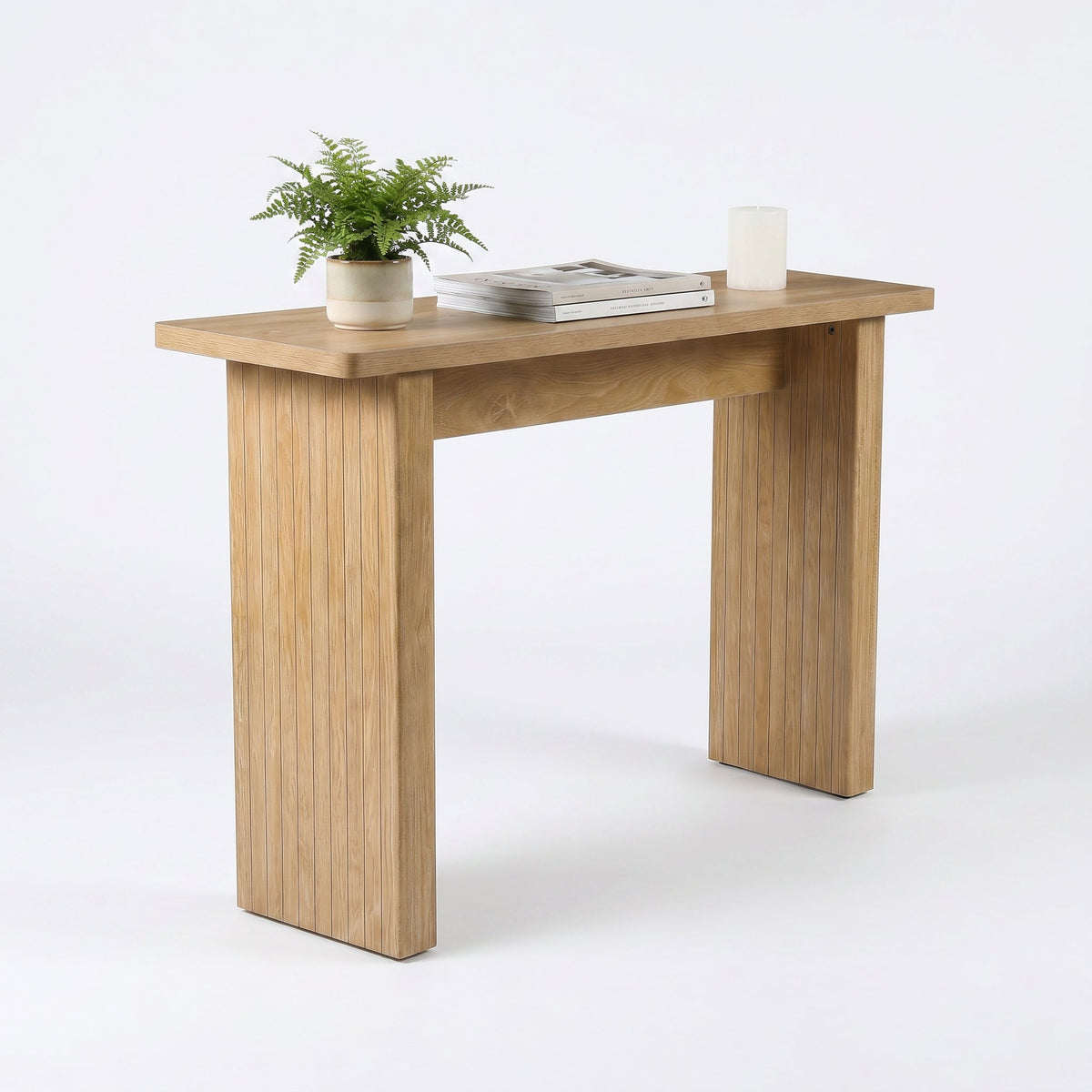 Light oak console table with vertical ribbed legs, styled with a potted fern, stacked books and white pillar candle, three…