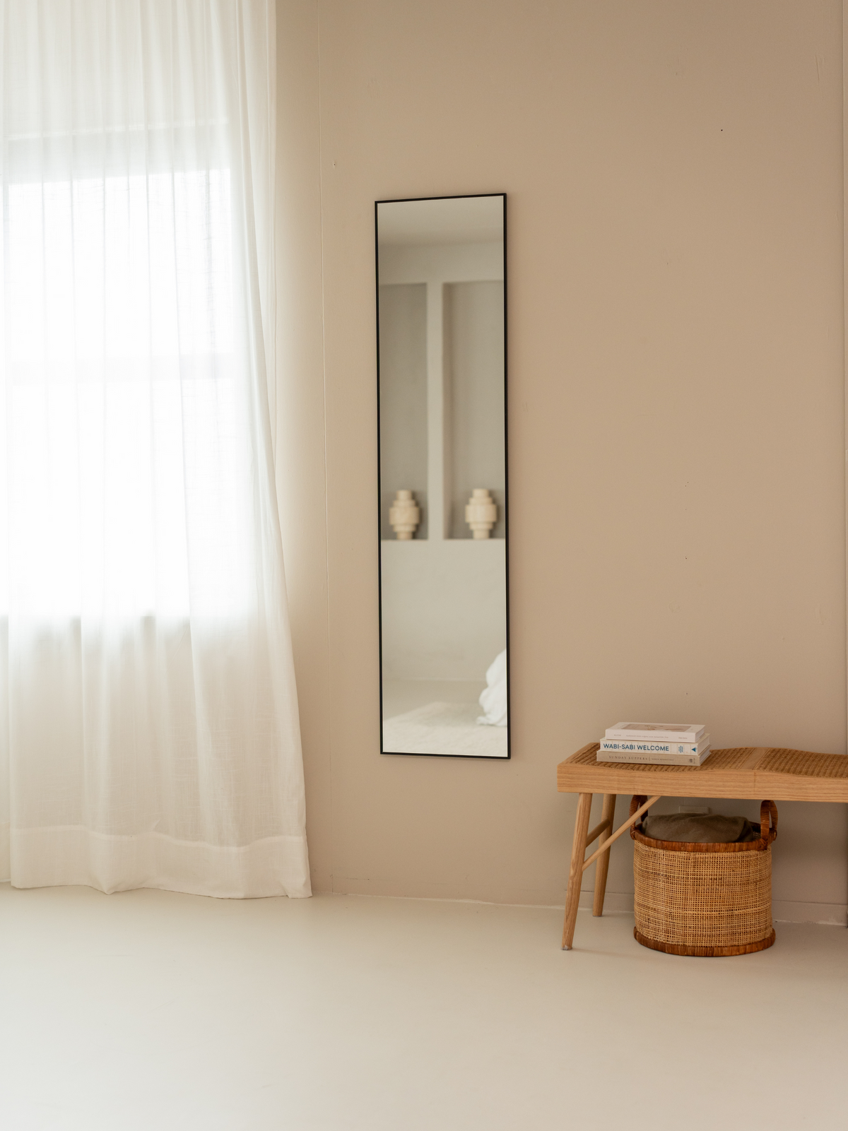 Slim black-framed full-length mirror wall-mounted in a warm beige room beside sheer curtains and a rattan bench with books