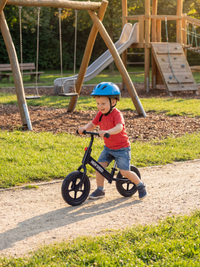 Toddler in blue helmet riding black New Speed balance bike along a playground path, swings and slide in background