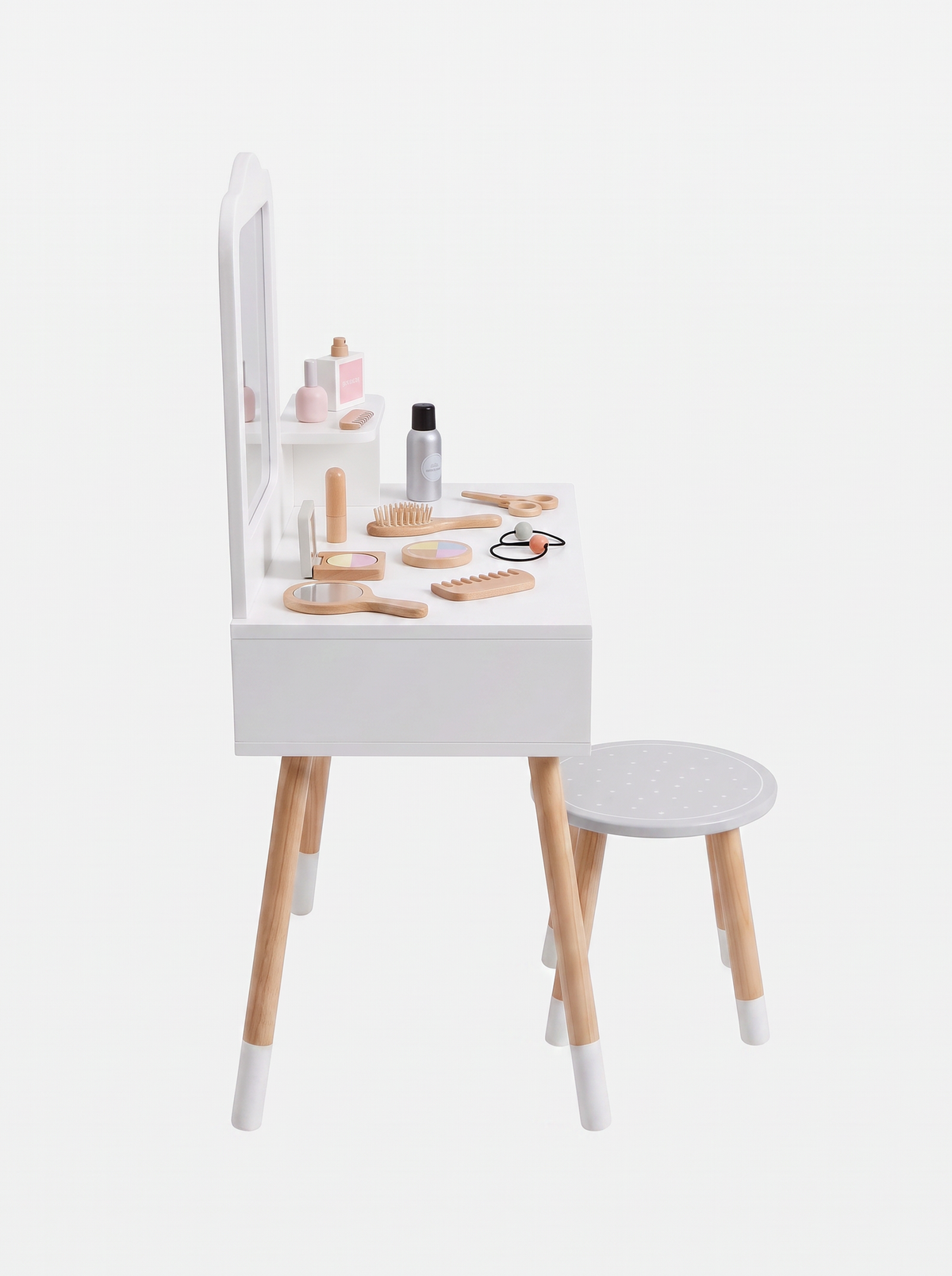 Angled side view of white wooden children's vanity with natural legs, mirror shelf, round grey stool, and scattered wooden…